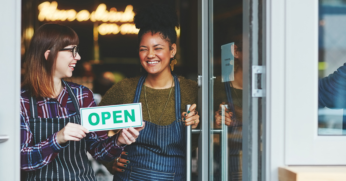 Two young female business owners stood smiling in doorway of business. One of them is holding an open sign